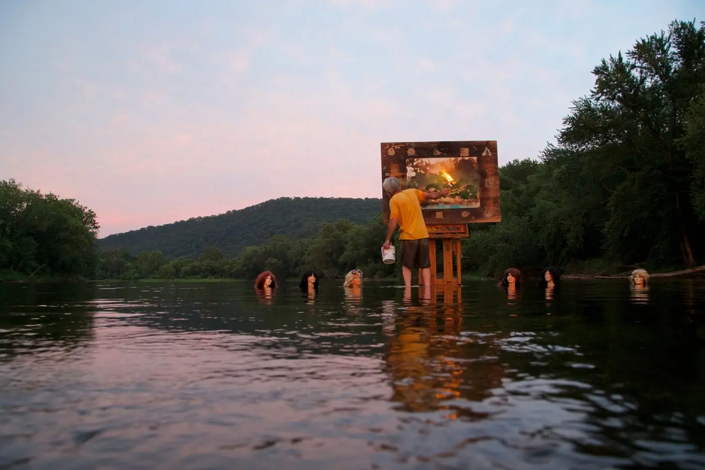 The artist painting a glowing fire scene at an easel standing in the Potomac River at sunset, watched by six mannequin heads in the water