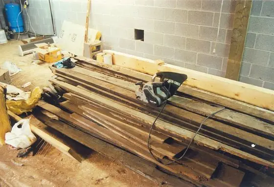 A stack of antique lumber on the basement floor with a belt sander on top, awaiting restoration