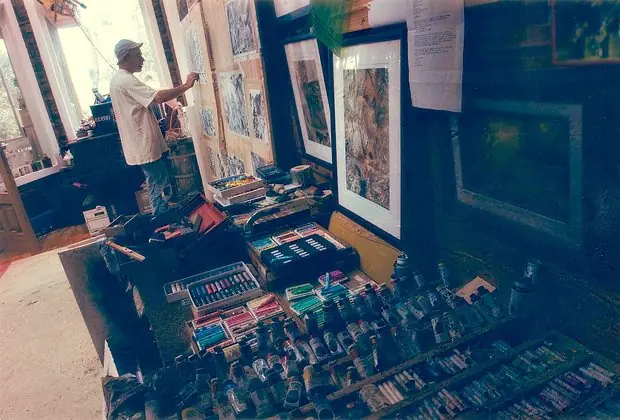 The artist drawing at a wall of charcoal pieces above a table of pastels and paint tubes