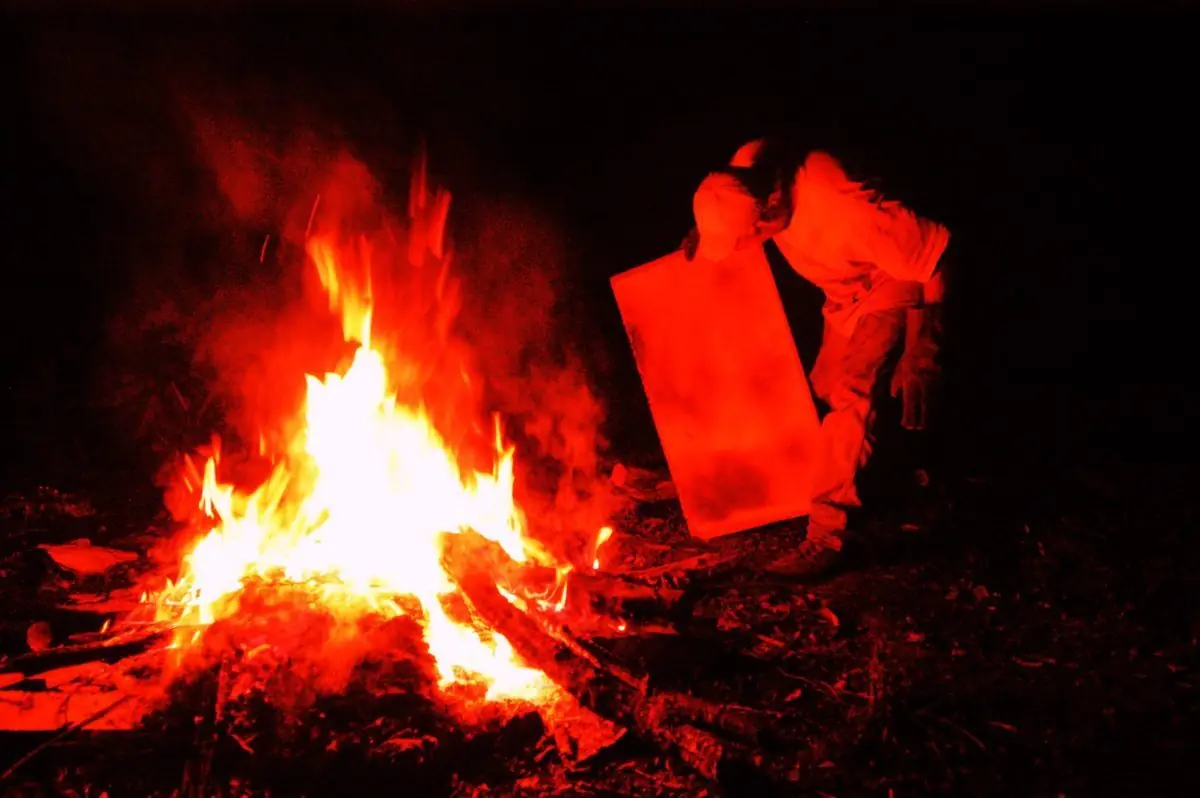 A figure leaning over a blazing bonfire at night, holding up a glowing red panel beside the flames