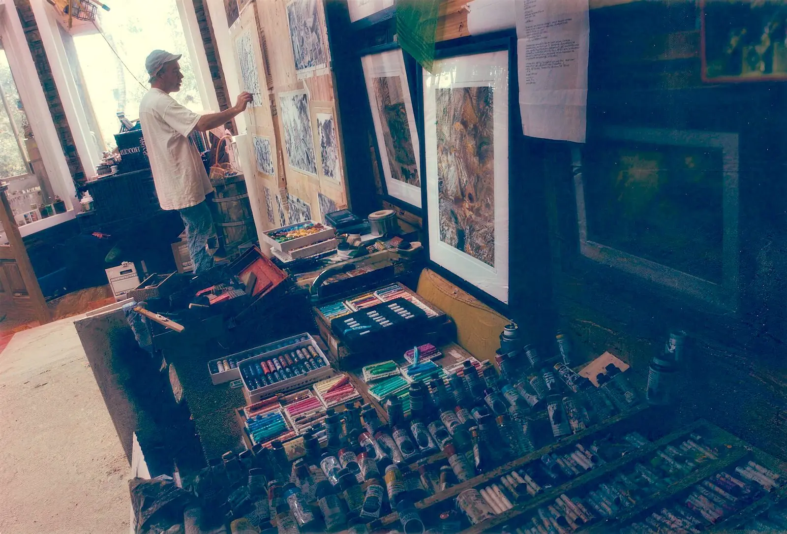 The artist drawing at a wall of charcoal pieces above a table of pastels and paint tubes