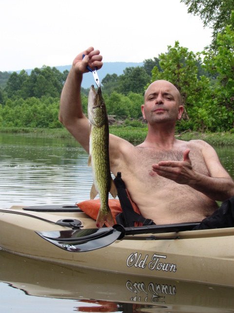 An angler holding up a pickerel, deadpan expression, river behind him.